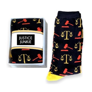 Pu&ograve; includere: Un paio di calzini neri con un motivo giallo e rosso di bilance della giustizia e simboli di martello. I calzini sono in una scatola bianca con un'etichetta che dice "Justice Junkie".