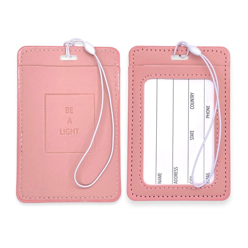 May include: Two pink leather luggage tags with white stitching. One tag has a white strap and the text "BE A LIGHT" embossed in a square. The other tag has a clear window for a name card and lines for writing contact information.