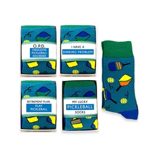 May include: Four boxes of pickleball themed socks with different sayings on each box. The sayings are "O.P.D. Obsessive Pickleball Disorder", "I Have a Dinkng Problem", "Retirement Plan Play Pickleball", and "My Lucky Pickleball Socks". There is also a single sock with a green cuff and a blue body with pickleball paddles and balls printed on it.