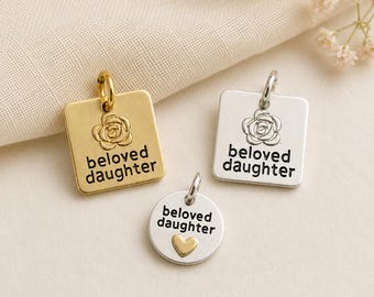 Könnte beinhalten: Zwei quadratische Metallanhänger in Gold- und Silbertönen, jeweils mit einer eingravierten Rose und dem Text "beloved daughter", sowie ein runder Silberanhänger mit dem Text "beloved daughter" und einem kleinen goldenen Herz, alle mit Ösen.