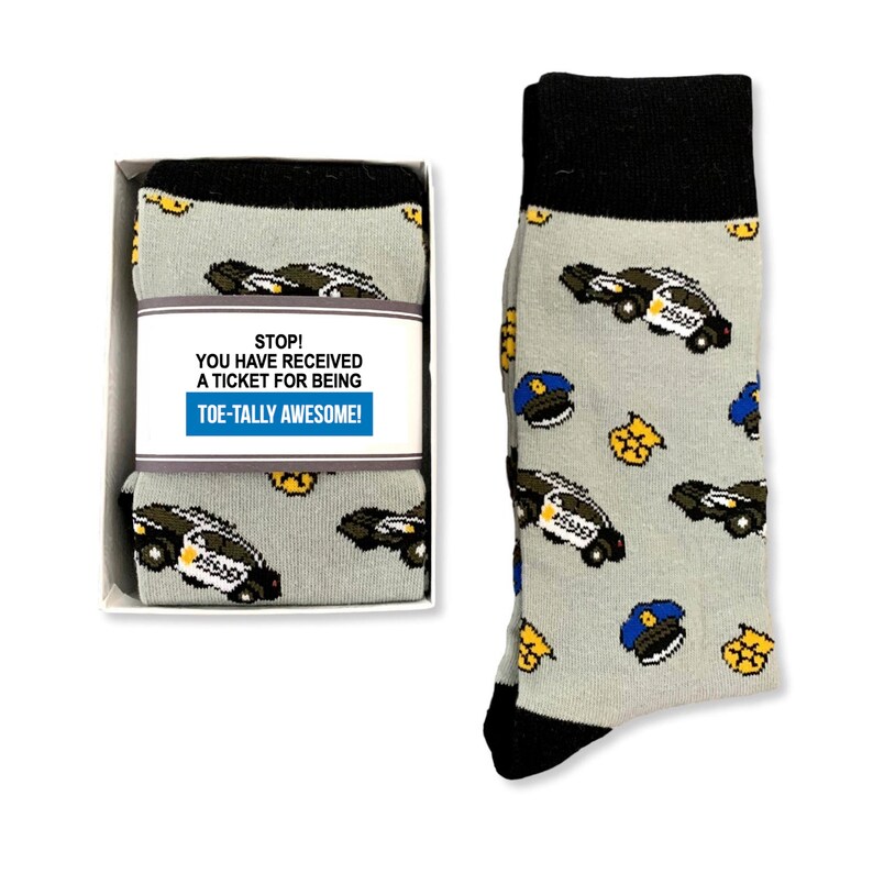Puede incluir: Calcetines grises con pu&ntilde;o y puntera negros, con un estampado de coches de polic&iacute;a, insignias y gorras. Los calcetines se presentan en una caja blanca con una etiqueta que dice: "STOP! YOU HAVE RECEIVED A TICKET FOR BEING TOE-TALLY AWESOME!"