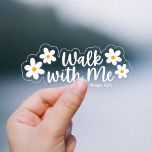 May include: Clear sticker with white text "Walk with Me" and the reference "Moses 6:34". The sticker is decorated with white daisy flowers with yellow centers. The sticker is held by a hand against a blurred background.