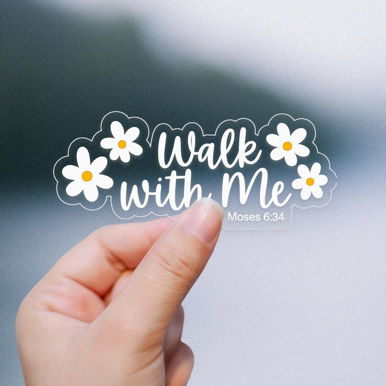 May include: Clear sticker with white text "Walk with Me" and the reference "Moses 6:34". The sticker is decorated with white daisy flowers with yellow centers. The sticker is held by a hand against a blurred background.
