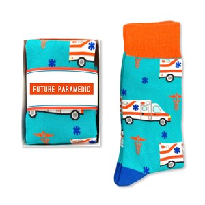 May include: A pair of blue socks with a pattern of orange and white ambulances and medical symbols. The socks are packaged in a white box with the text "FUTURE PARAMEDIC" on the label.
