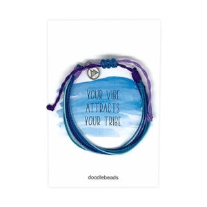 May include: A multi-colored friendship bracelet with a silver charm that says "Your vibe attracts your tribe". The bracelet is on a white card with the words "doodlebeads" printed on it.