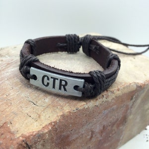 Op de afbeelding: Bruin leren armband met een zilveren metalen tag waarop "CTR" staat.