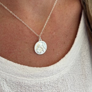 May include: A silver chain necklace with a round pendant featuring a detailed embossed design. The pendant is slightly textured.
