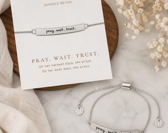 May include: A silver-toned adjustable bracelet featuring a curved metal bar engraved with the text pray. wait. trust. The bracelet is displayed on a white card with the brand name DOODLE BEADS and the printed text PRAY. WAIT. TRUST. On her hardest days, she prays. On her best days, she prays.