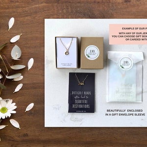 Pode incluir: Um colar de ouro com um pequeno pingente de montanha, embalado em uma caixa de presente branca com um cart&atilde;o preto que diz "Difficult roads often lead to beautiful destinations". O colar tamb&eacute;m &eacute; mostrado em um envelope de pl&aacute;stico transparente com um cart&atilde;o branco que diz "Beautifully enclosed in a gift envelope sleeve".