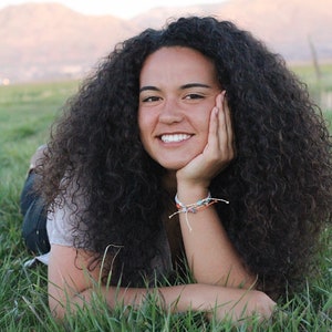 May include: A young woman with long, curly dark brown hair is lying in a field of green grass. She is smiling and has her hand on her cheek. She is wearing a light gray shirt and a bracelet on her wrist.
