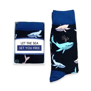 May include: A pair of black socks with a blue cuff and toe, featuring a pattern of light blue and pink whales. The socks are displayed next to a box with the text "LET THE SEA SET YOU FREE".