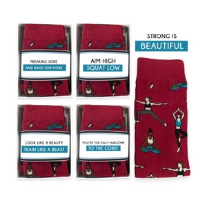 Könnte beinhalten: Vier Paar rote Socken mit weißem Besatz und verschiedenen Sprüchen zum Thema Yoga. Die Socken zeigen ein Muster von Frauen in verschiedenen Yoga-Posen. Die Socken sind in weißen Schachteln verpackt, auf denen die Sprüche aufgedruckt sind. Zu den Socken gehört eine größere rote Socke mit dem gleichen Muster und dem Text "Strong is Beautiful".