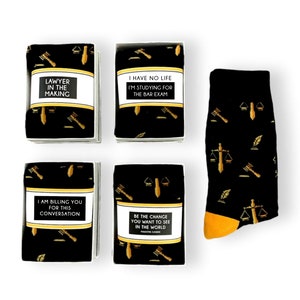 May include: Four black gift boxes with white trim and gold text on the front. The boxes contain socks with a black and gold pattern of scales of justice, gavel, and quill. The text on the boxes reads: "Lawyer in the Making", "I have no life I'm studying for the bar exam", "I am billing you for this conversation", and "Be the change you want to see in the world Mahatma Gandhi".