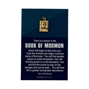May include: A gold charm shaped like a book with the words "The Book of Mormon" on the cover. The charm is on a blue background with a quote from Ezra Taft Benson about the power of the Book of Mormon. The quote reads: "There is a power in the Book of Mormon which will begin to flow into your lives the moment you begin a serious study of the book. You will find greater power to resist temptation. You will find the power to avoid deception. You will find the power to stay on the straight and narrow path... You will find life in greater and greater abundance." -Ezra Taft Benson. The background also includes the words "Doodle Beads".