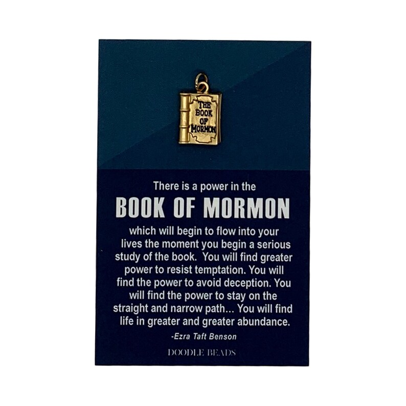 May include: A gold charm shaped like a book with the words "The Book of Mormon" on the cover. The charm is on a blue background with a quote from Ezra Taft Benson about the power of the Book of Mormon. The quote reads: "There is a power in the Book of Mormon which will begin to flow into your lives the moment you begin a serious study of the book. You will find greater power to resist temptation. You will find the power to avoid deception. You will find the power to stay on the straight and narrow path... You will find life in greater and greater abundance." -Ezra Taft Benson. The background also includes the words "Doodle Beads".
