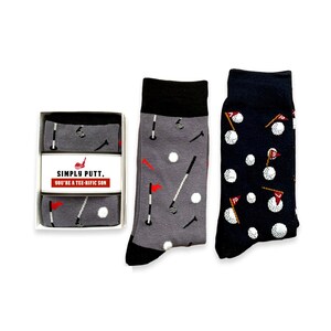 May include: Two pairs of socks with a golf theme. One pair is gray with white golf balls, golf clubs, and flags. The other pair is navy blue with white golf balls and flags. The socks come in a box with the text "Simply Putt. You're a Tee-Rific Son."