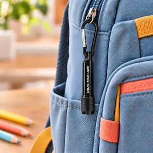 May include: A small black cylindrical flashlight with a textured grip and the text SHARE YOUR LIGHT printed in white, attached to a silver metal carabiner clip hanging from the blue fabric zipper pull of a blue and orange backpack.