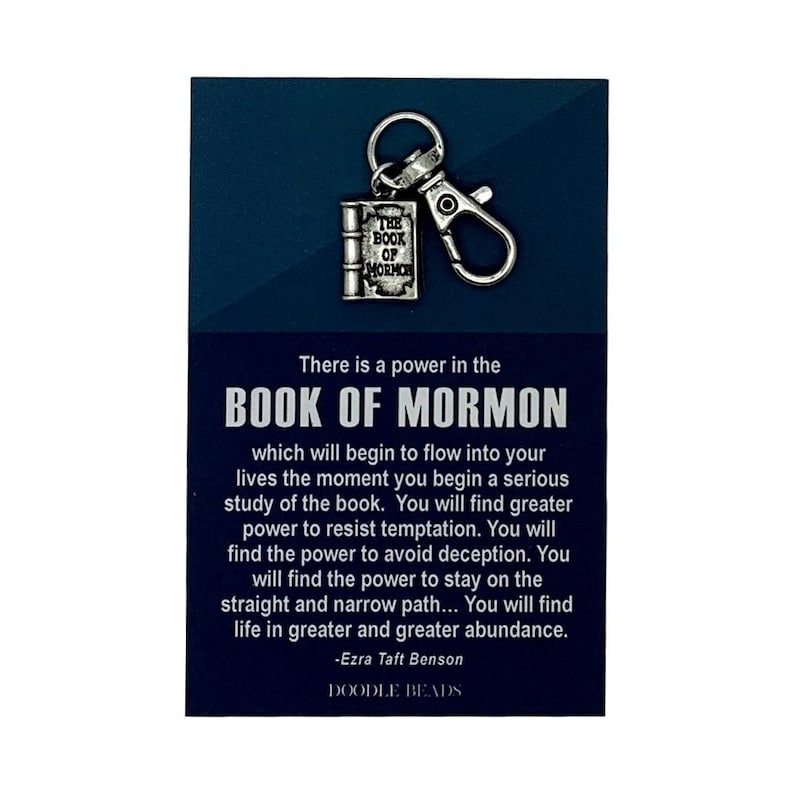 May include: A silver keychain charm shaped like a closed book with the words "The Book of Mormon" on the cover. The charm is attached to a silver split ring and a silver lobster clasp. The background is a dark blue card with the text "There is a power in the Book of Mormon which will begin to flow into your lives the moment you begin a serious study of the book. You will find greater power to resist temptation. You will find the power to avoid deception. You will find the power to stay on the straight and narrow path... You will find life in greater and greater abundance. -Ezra Taft Benson DOODLE BEADS"