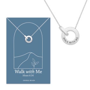 Pode incluir: Um colar de prata com um pingente circular gravado com as palavras "Walk with Me". O pingente está conectado a uma corrente delicada. O colar é exibido em um cartão azul com o texto "Walk with Me" e a referência bíblica "Moisés 6:34".