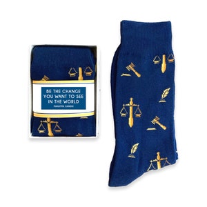 May include: A pair of blue socks with a gold pattern of scales of justice, gavel, and quill pens. The socks are in a white box with a quote from Mahatma Gandhi: "Be the change you want to see in the world."