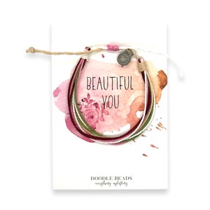 May include: A multi-colored string bracelet with a small silver heart charm. The bracelet is displayed on a white card with a watercolor background and the text "Beautiful You" in black lettering. The card also includes the text "Doodle Beads everything uplifting".