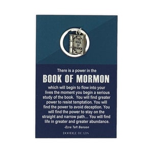 May include: A silver charm keychain featuring a closed book with the words "The Book of Mormon" on the cover. The charm is attached to a silver key ring. The keychain is displayed on a blue card with a quote about the power of the Book of Mormon by Ezra Taft Benson. The card also includes the text "Doodle Beads".