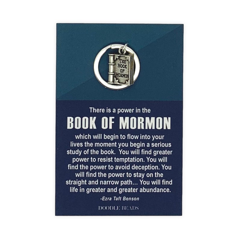 May include: A silver charm keychain featuring a closed book with the words "The Book of Mormon" on the cover. The charm is attached to a silver key ring. The keychain is displayed on a blue card with a quote about the power of the Book of Mormon by Ezra Taft Benson. The card also includes the text "Doodle Beads".