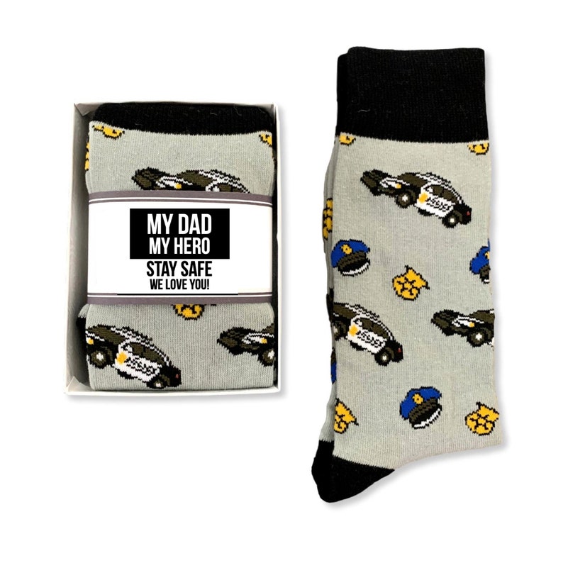 Puede incluir: Calcetines grises con pu&ntilde;o y puntera negros, con un estampado de coches de polic&iacute;a, insignias y gorras. Los calcetines est&aacute;n en una caja blanca con la etiqueta "MY DAD MY HERO STAY SAFE WE LOVE YOU!"