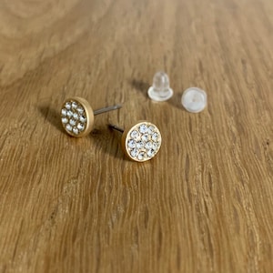 May include: A pair of gold stud earrings with clear crystal accents. The earrings are on a wooden surface with two clear plastic earring backs.