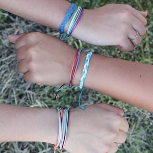 May include: Two wrists with colorful friendship bracelets. The bracelets are made of woven string in various colors, including blue, green, red, orange, and white.
