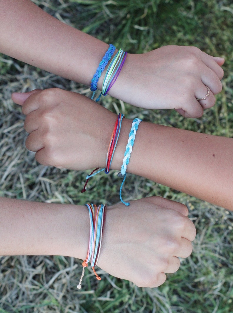 May include: Two wrists with colorful friendship bracelets. The bracelets are made of woven string in various colors, including blue, green, red, orange, and white.
