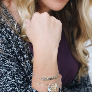 May include: A silver bangle bracelet with a charm that says "begin today" and a clear crystal charm. The bracelet is on a woman's wrist.