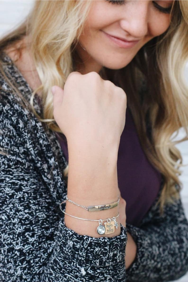 May include: A silver bangle bracelet with a charm that says "begin today" and a clear crystal charm. The bracelet is on a woman's wrist.