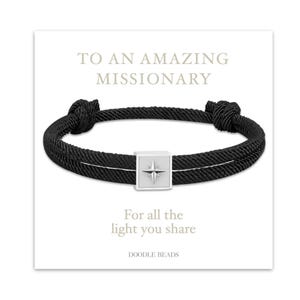 May include: Black rope bracelet with a silver square centerpiece featuring a compass rose design. The bracelet is displayed on a white card with the text "TO AN AMAZING MISSIONARY" and "For all the light you share."