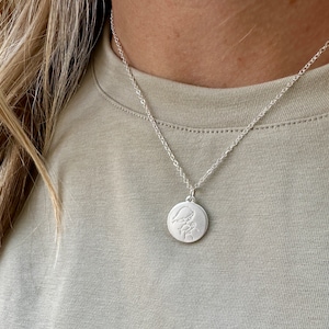 May include: A silver necklace with a round pendant featuring a delicate engraved design. The pendant is on a silver chain.