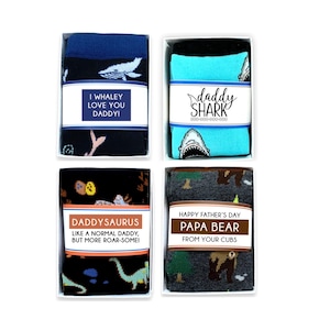 May include: Four gift boxes containing pairs of socks with different designs and messages. The socks are black with a whale, blue with a shark, black with dinosaurs, and gray with bears. The messages on the boxes are "I Whaley Love You Daddy!", "Daddy Shark", "Daddysaurus Like a Normal Daddy, But More Roar-Some!", and "Happy Father's Day Papa Bear From Your Cubs".