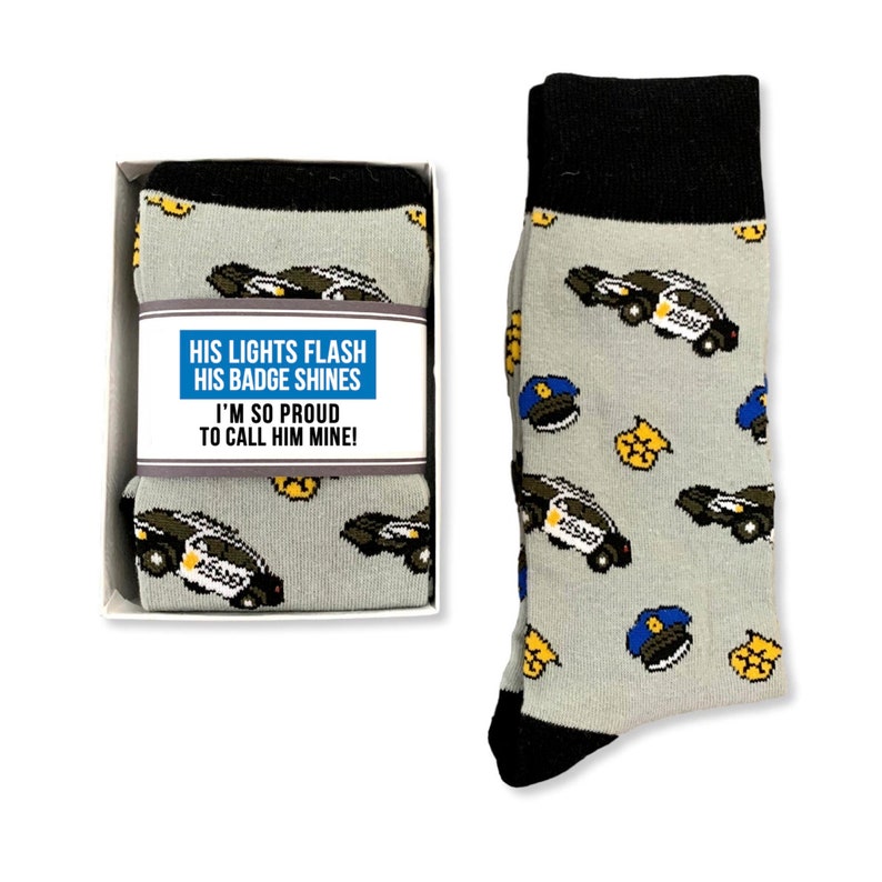 Puede incluir: Calcetines grises con pu&ntilde;o y puntera negros, con un estampado de coches de polic&iacute;a, insignias y gorras. Los calcetines se presentan con una caja de regalo que lleva el texto "HIS LIGHTS FLASH HIS BADGE SHINES I'M SO PROUD TO CALL HIM MINE!"