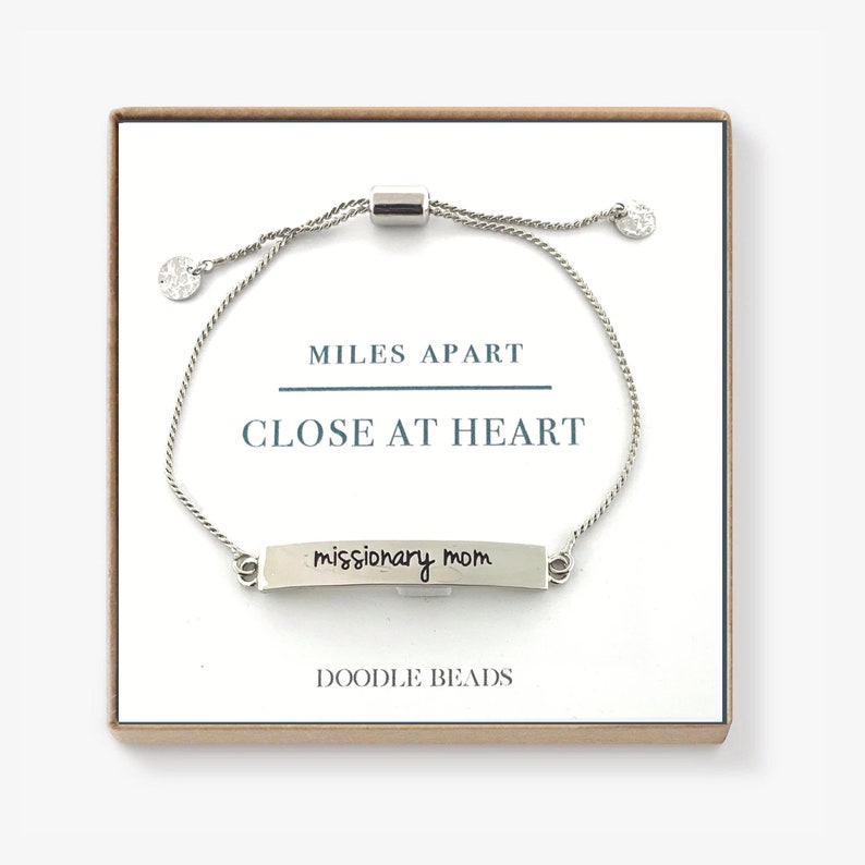 May include: Silver bracelet with a silver charm that says "missionary mom" and a silver slider bead. The bracelet is in a box with the words "Miles Apart Close At Heart" and "Doodle Beads" printed on it.