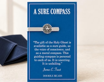May include: A silver-toned tie bar featuring a circular compass emblem attached to a blue display card. The card displays the text A SURE COMPASS and a quote by James E. Faust about the Holy Ghost, ending with the brand name DOODLE BEADS.