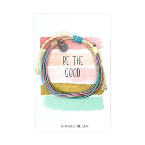 May include: A colorful friendship bracelet with a silver heart charm and the words "BE THE GOOD" on a white card with a watercolor background.