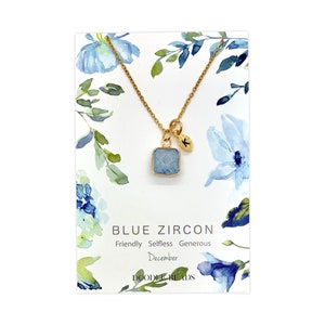Può includere: Collana d'oro con un ciondolo quadrato blu e un piccolo ciondolo a forma di foglia d'oro. La carta recita "Blue Zircon, Friendly, Selfless, Generous, December, Doodle Beads".