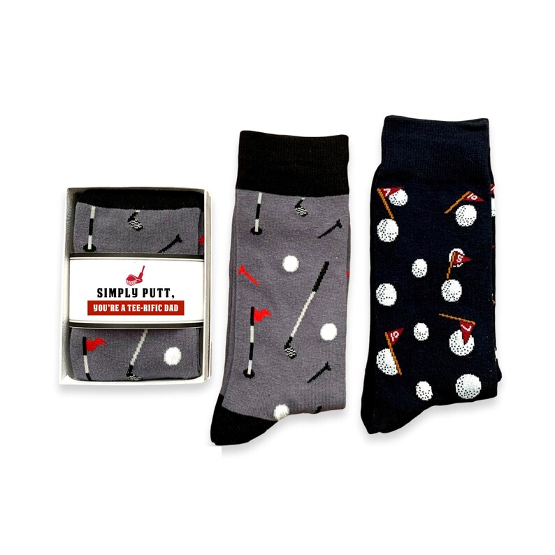 May include: A pair of gray and navy blue socks with a golf theme. The socks feature golf clubs, golf balls, and flags. The gray socks have the text "Simply Putt, You're a Tee-Rific Dad".
