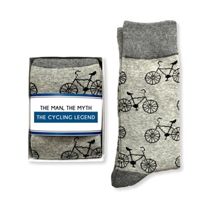 May include: A gift box containing a pair of gray socks with a black bicycle pattern. The box has a label that reads "The Man, The Myth, The Cycling Legend."