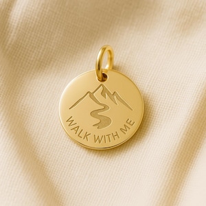 Op de afbeelding: Goudkleurige hanger met een berg- en rivierontwerp. De hanger heeft de inscriptie "WALK WITH ME" rondom. Het is rond met een kleine lus aan de bovenkant voor een ketting.