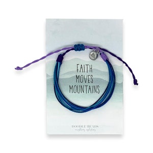 Puede incluir: Un brazalete de cuerda multicolor con un dije plateado que dice "Faith Moves Mountains". El brazalete está sobre una tarjeta blanca con las palabras "Doodle Beads" y "everything meaningful" impresas en ella.