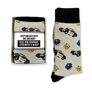 Puede incluir: Calcetines grises con pu&ntilde;o y puntera negros, con un estampado de coches de polic&iacute;a, insignias y gorras. Los calcetines se muestran junto a un par en caja con la etiqueta "KEEP HIM SAFE BOTH DAY AND NIGHT. GIVE HIM COURAGE, STRENGTH & MIGHT."
