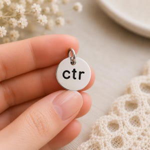 Könnte beinhalten: Ein kleiner, kreisförmiger Anhänger aus silberfarbenem Metall mit den in Schwarz eingravierten Kleinbuchstaben "ctr". Der Anhänger verfügt über einen kleinen Biegering an der Oberseite zur Befestigung und hat eine polierte Oberfläche.