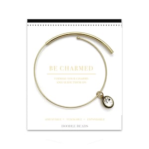 May include: Gold bangle bracelet with a clear crystal charm. The bracelet is adjustable, stackable, and expandable. The text "BE CHARMED" and "DOODLE BEADS" is printed on the card.
