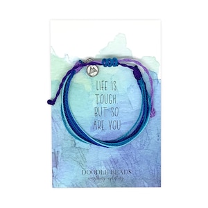 May include: A multi-colored string bracelet with a silver charm that says "Life is tough, but so are you". The bracelet is on a card with a watercolor background and the words "Doodle Beads, everything uplifting".
