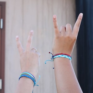 May include: Two hands with bracelets on each wrist. The hand on the left is making a peace sign, while the hand on the right is making a rock and roll sign. The bracelets are made of colorful string.